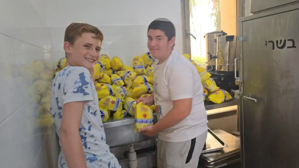 שני תלמידים מבית הספר אמית בר אילן פתח תקווה מחייכים בזמן שהם אורזים עופות לחלוקת מזון למשפחות נזקקות לקראת החג.