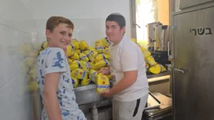 שני תלמידים מבית הספר אמית בר אילן פתח תקווה מחייכים בזמן שהם אורזים עופות לחלוקת מזון למשפחות נזקקות לקראת החג.