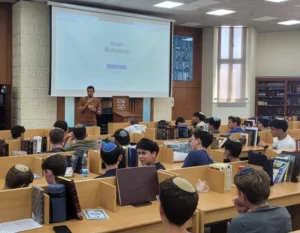 תלמידי ישיבת אמית כפר גנים בפתח תקווה יושבים בבית המדרש במהלך השקת תוכנית המדריכים, כשאחד המדריכים מציג את התוכנית.