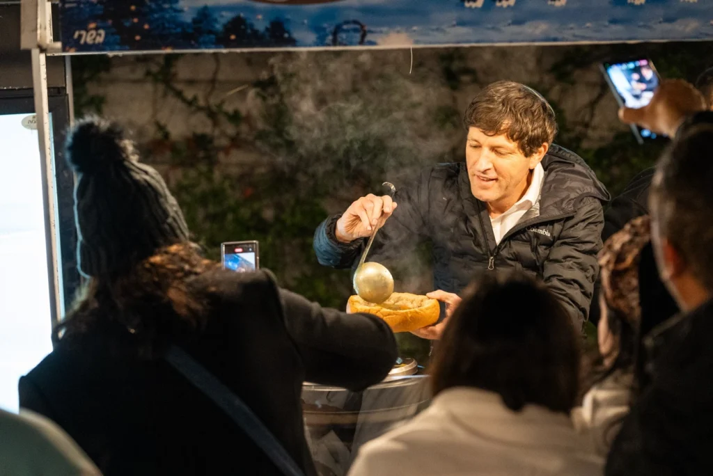 ראש העיר פתח תקווה, רמי גרינברג, מחייך ומוזג מרק חם מתוך קערת לחם גדולה בדוכן אוכל בפסטיבל לילי.